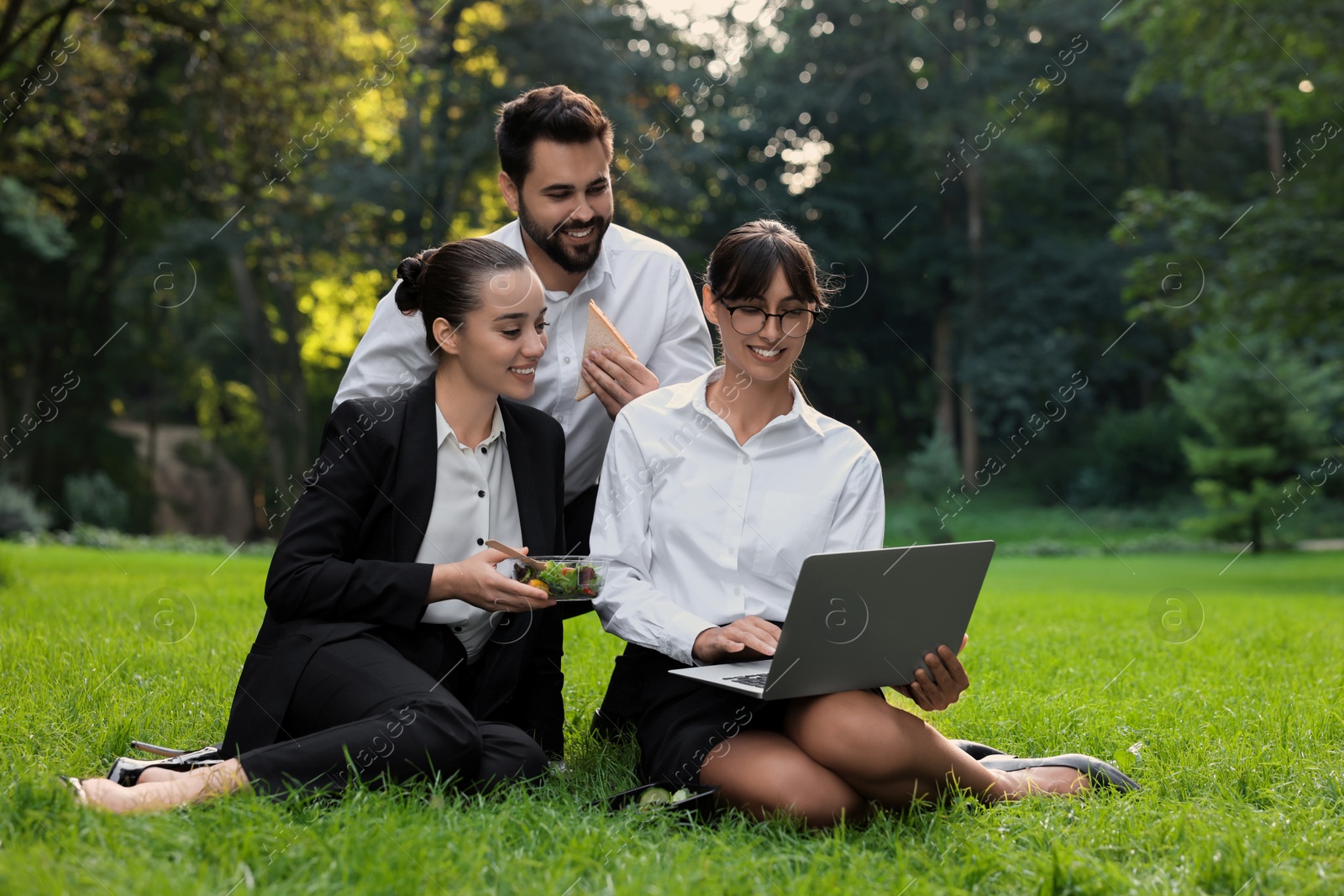 Photo of Happy colleagues with laptop having business lunch on green grass in park