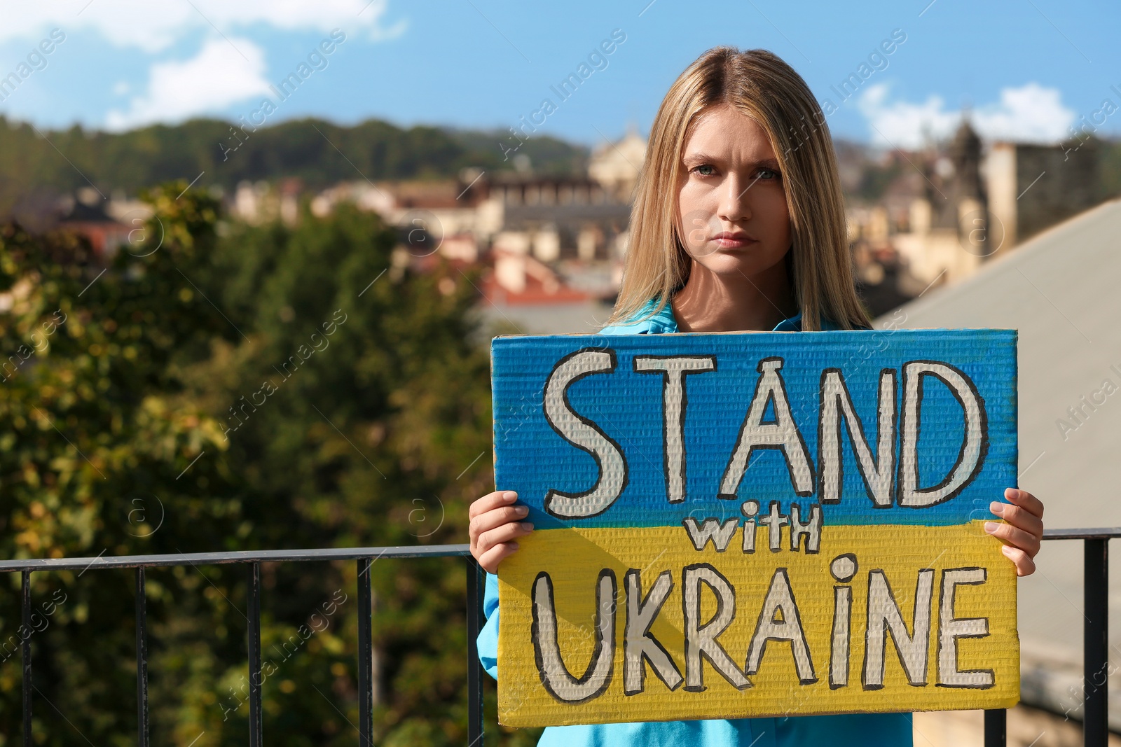 Photo of Sad woman holding poster in colors of national flag and words Stand with Ukraine outdoors. Space for text