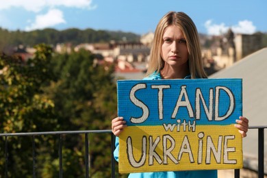 Photo of Sad woman holding poster in colors of national flag and words Stand with Ukraine outdoors. Space for text