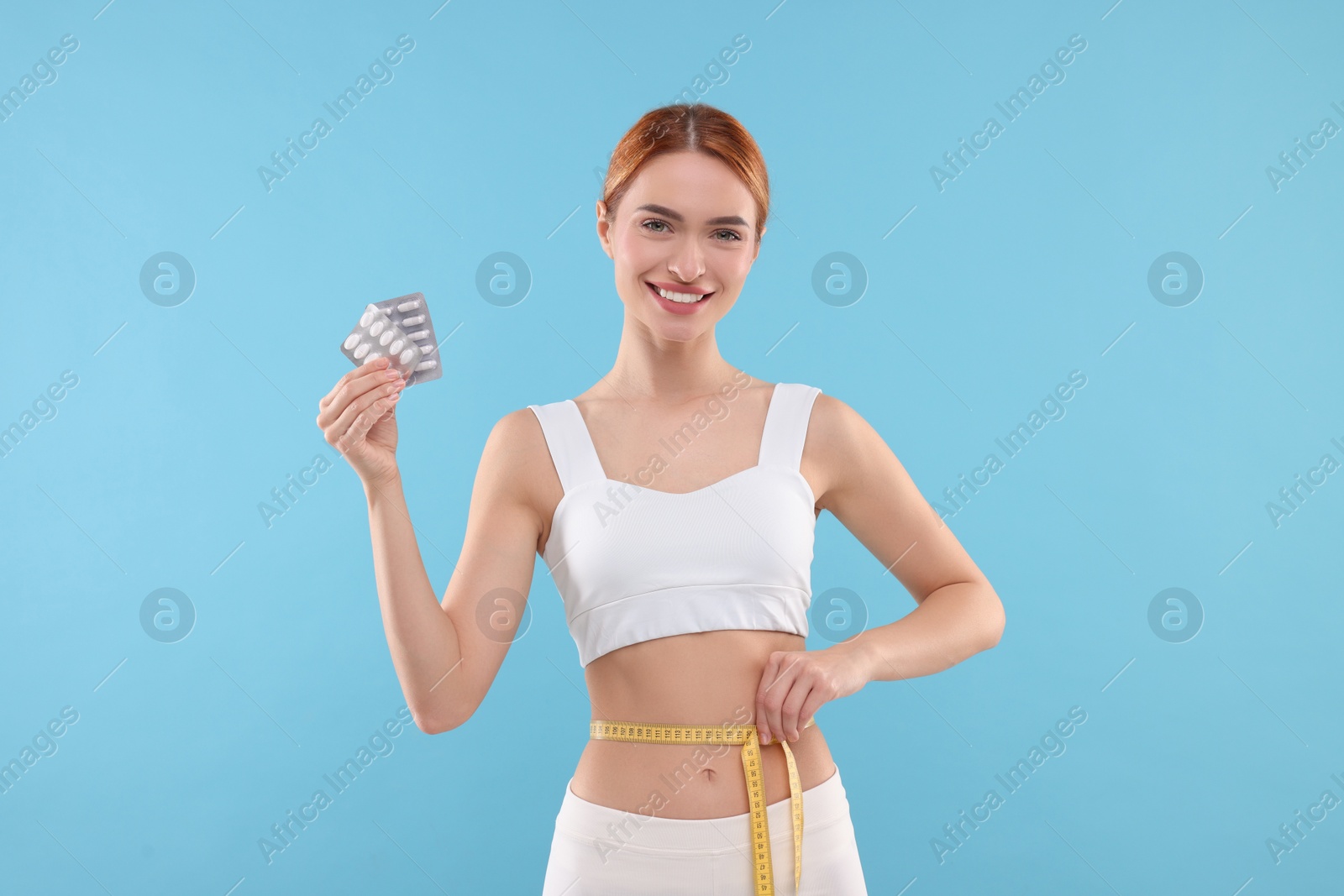 Photo of Slim woman holding pills and measuring waist with tape on light blue background. Weight loss