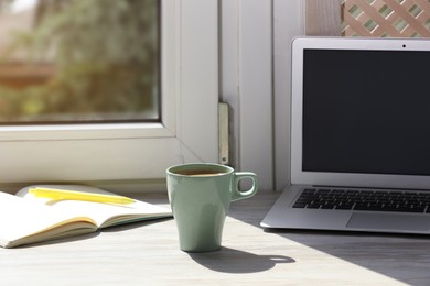 Photo of Cup of coffee, laptop with blank screen, stationery on wooden window sill. Space for text