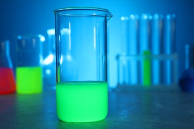 Photo of Laboratory beaker with luminous liquid on table against blurred background, closeup