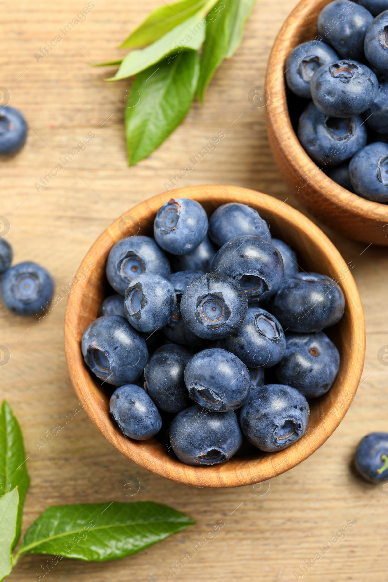 Photo of Bowls of fresh tasty blueberries and leaves on wooden table, flat lay