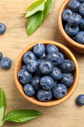 Photo of Bowls of fresh tasty blueberries and leaves on wooden table, flat lay