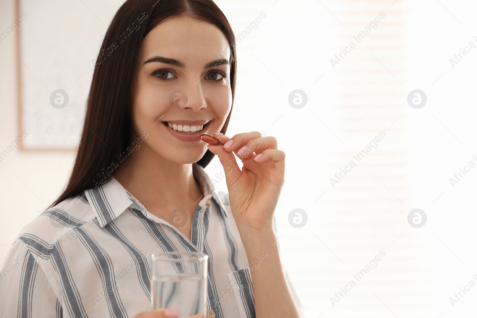 Photo of Young woman with glass of water taking vitamin capsule indoors. Space for text