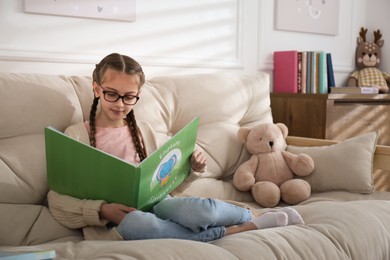 Cute little girl reading book on sofa at home