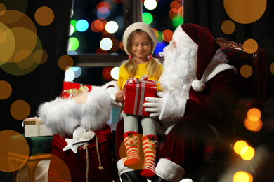 Photo of Santa Claus and little girl with Christmas gift near window indoors