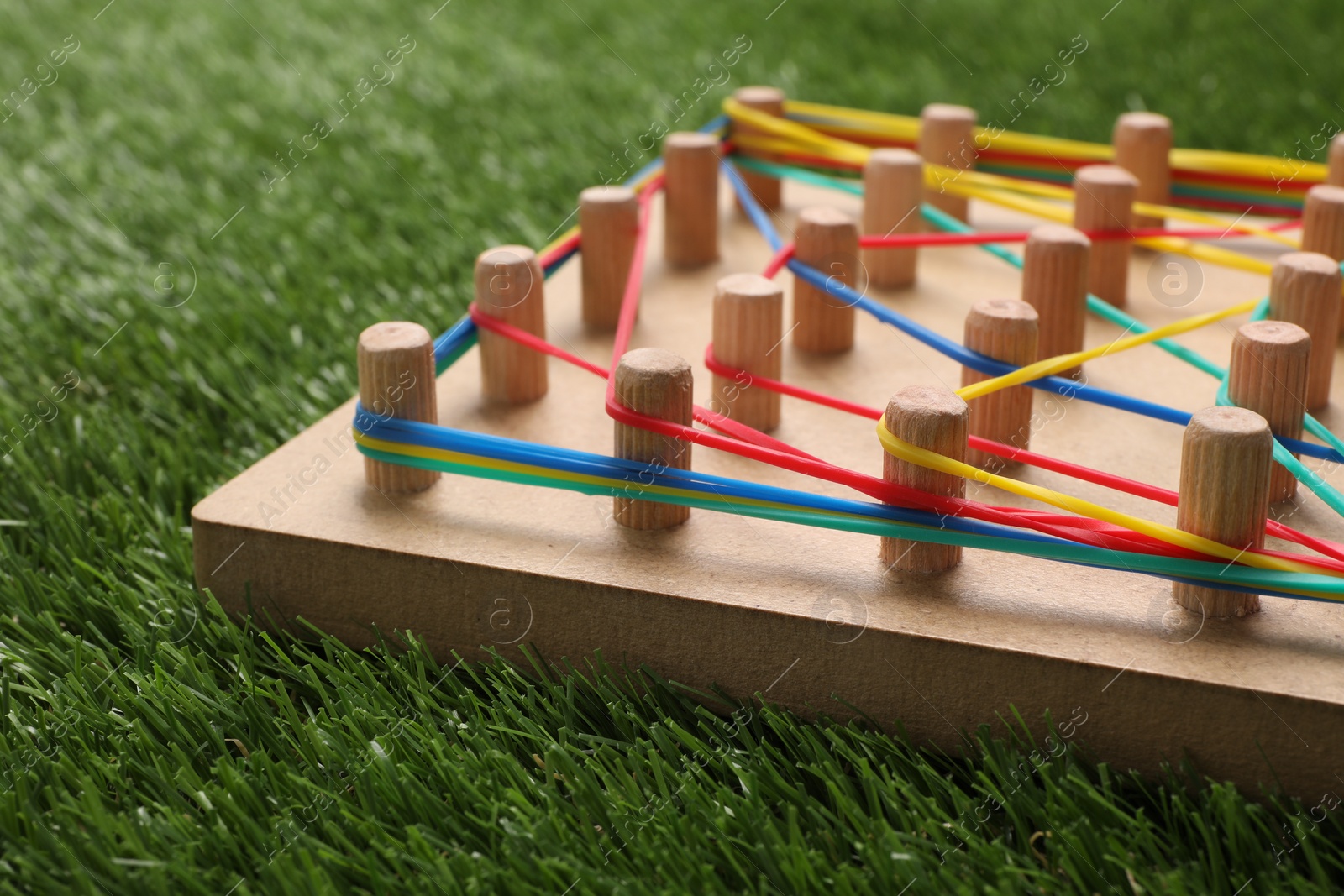 Photo of Wooden geoboard with rubber bands on artificial grass, closeup. Educational toy for motor skills development