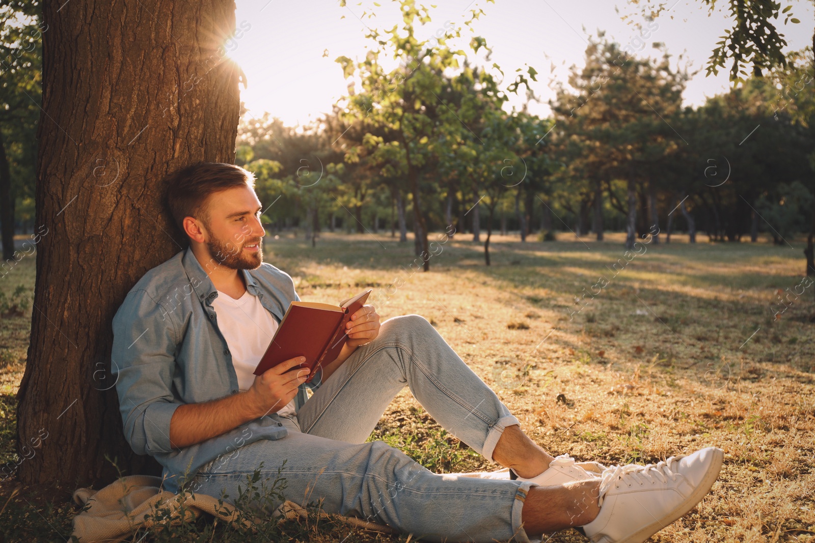 Photo of Young man reading book on green grass near tree in park