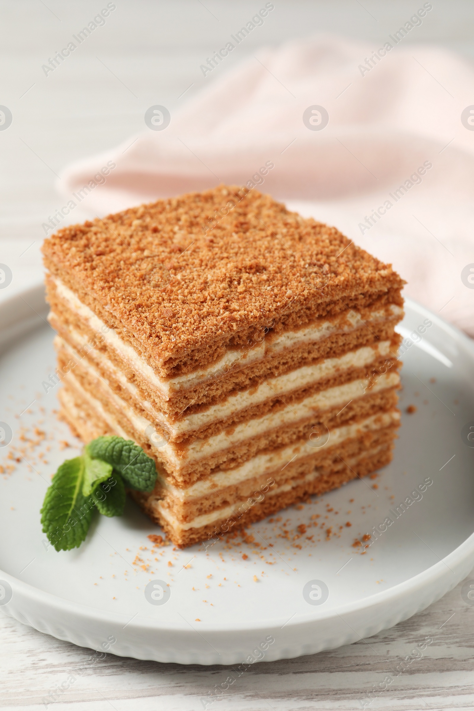 Photo of Slice of delicious layered honey cake with mint served on white wooden table, closeup