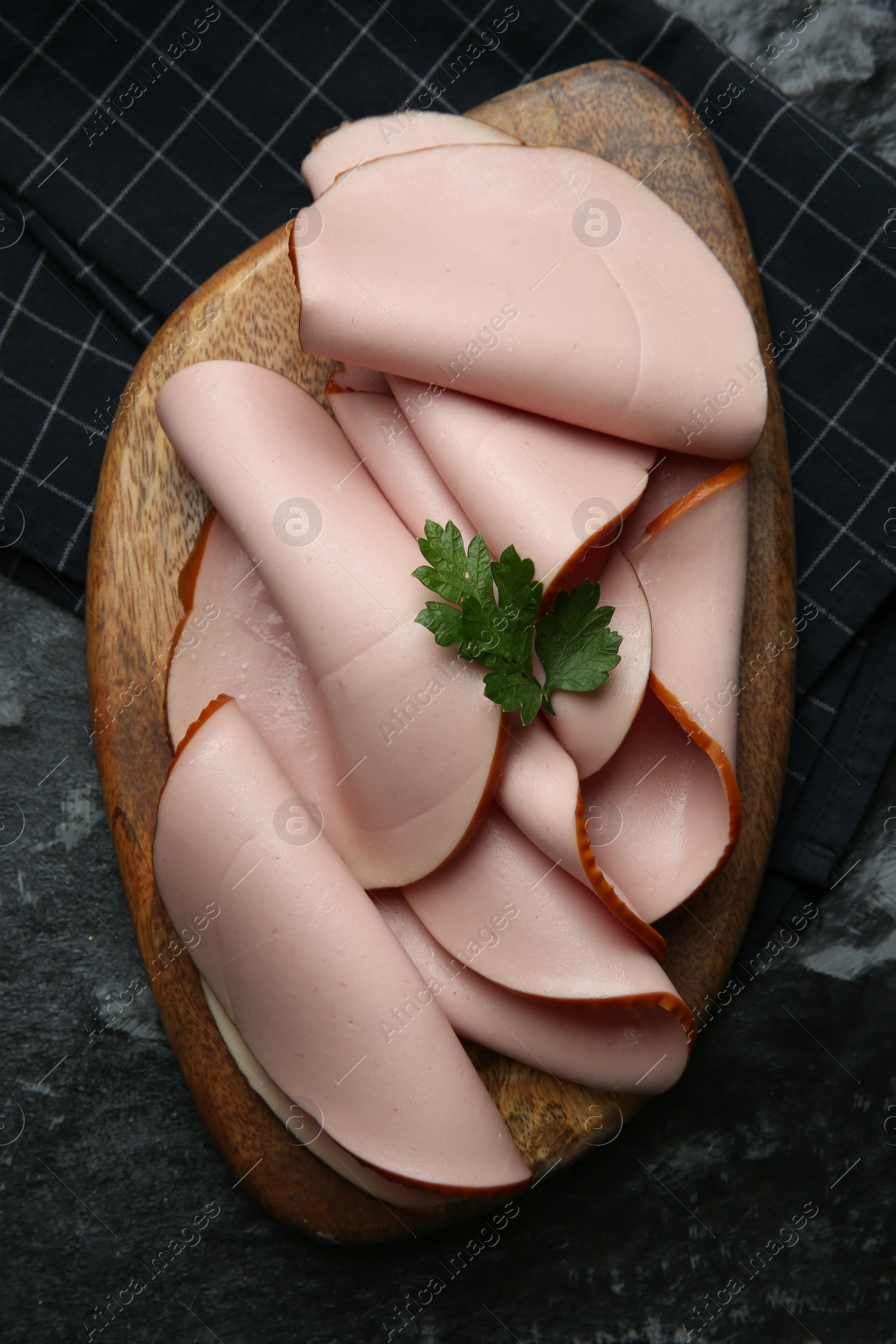 Photo of Board with slices of tasty boiled sausage and parsley on dark textured table, top view