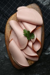 Board with slices of tasty boiled sausage and parsley on dark textured table, top view