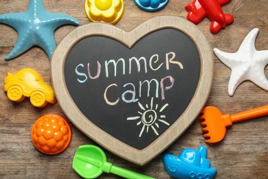 Photo of Heart shaped chalkboard with text SUMMER CAMP and different sand molds on wooden background, flat lay