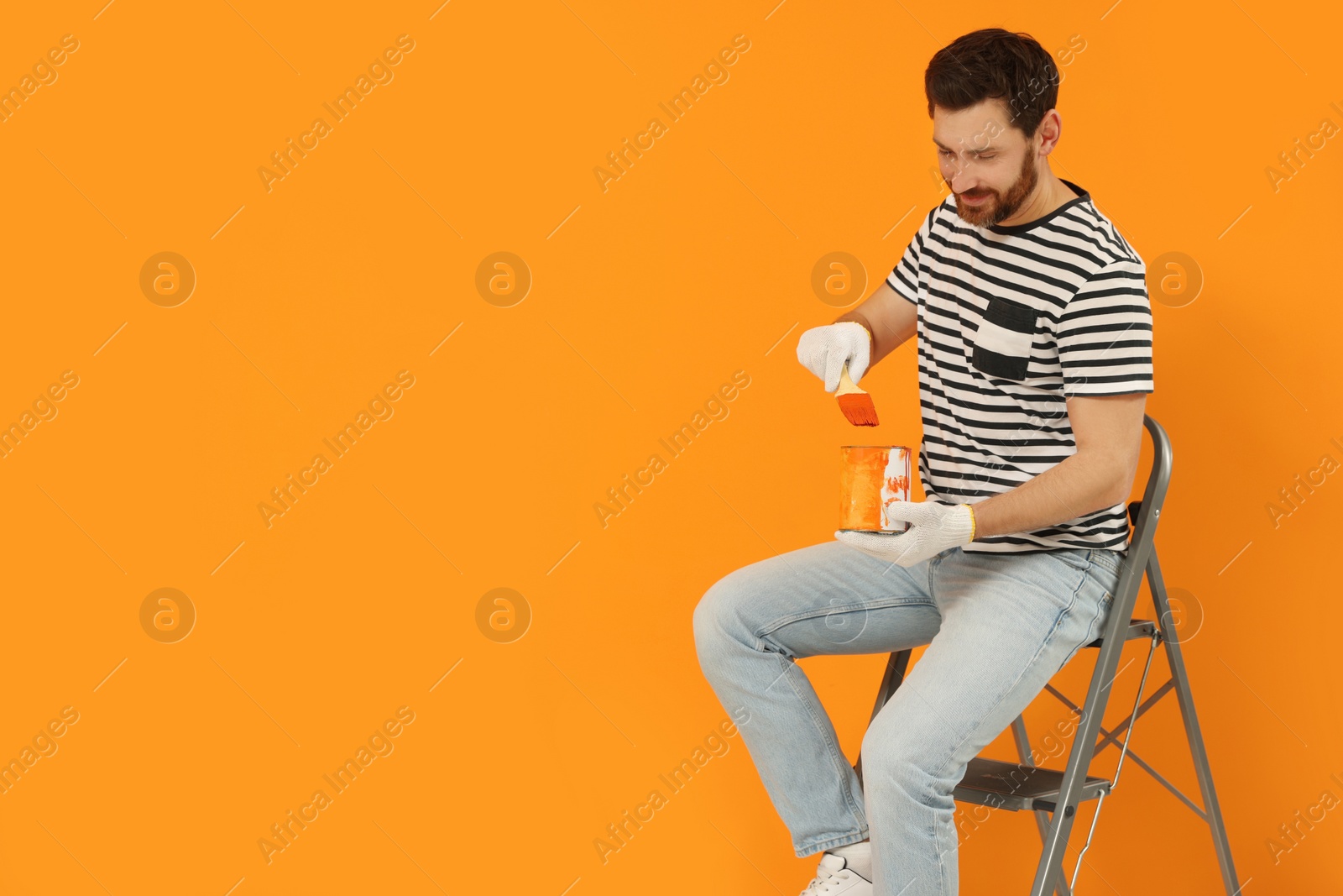 Photo of Designer with painting equipment sitting on folding ladder near freshly painted orange wall, space for text