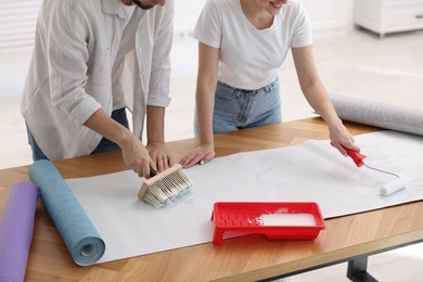 Woman and man applying glue onto wallpaper sheet at wooden table indoors, closeup