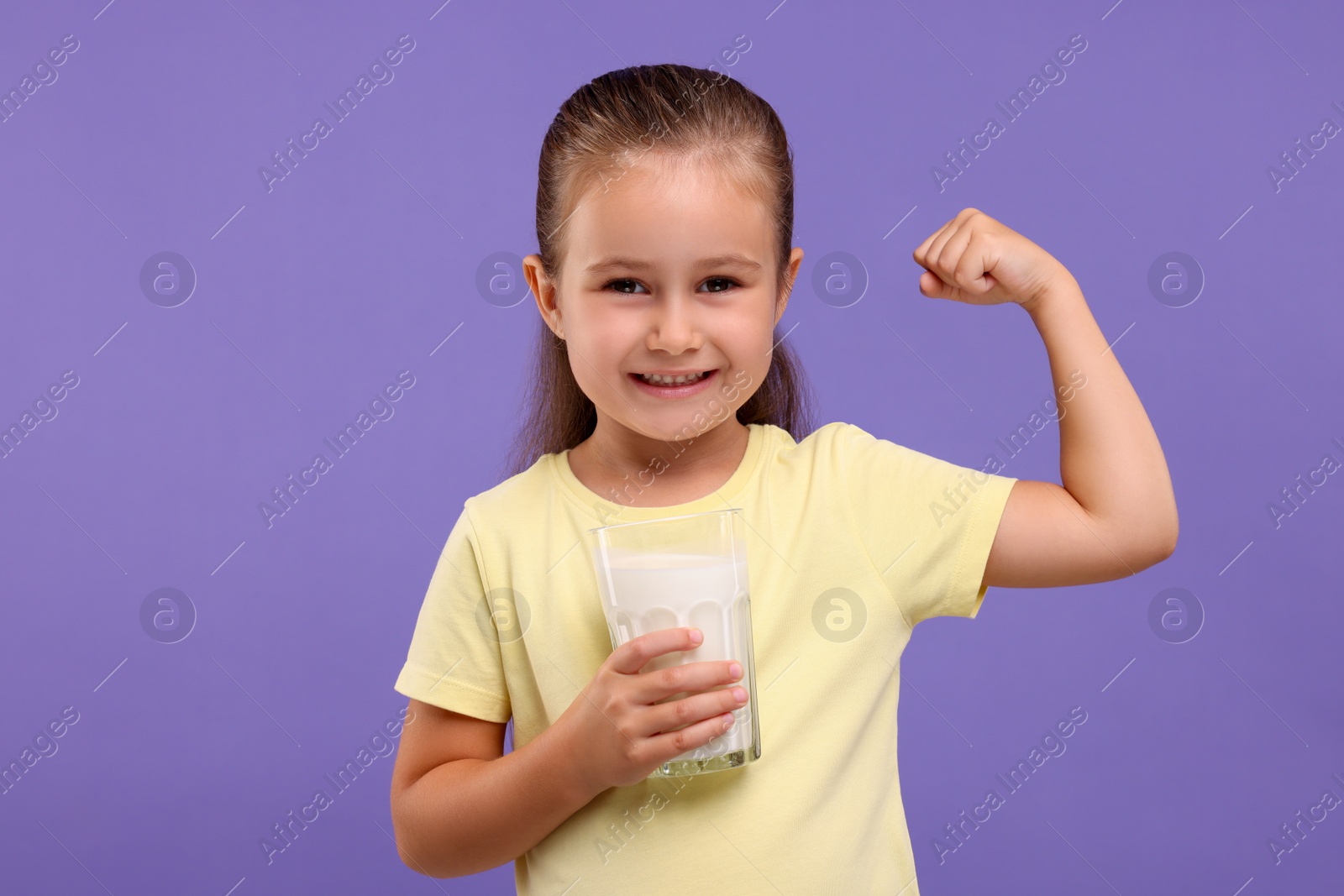 Photo of Cute girl with glass of fresh milk showing her strength on violet background