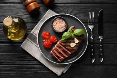 Photo of Delicious grilled beef steak served with spices and tomatoes on black wooden table, flat lay