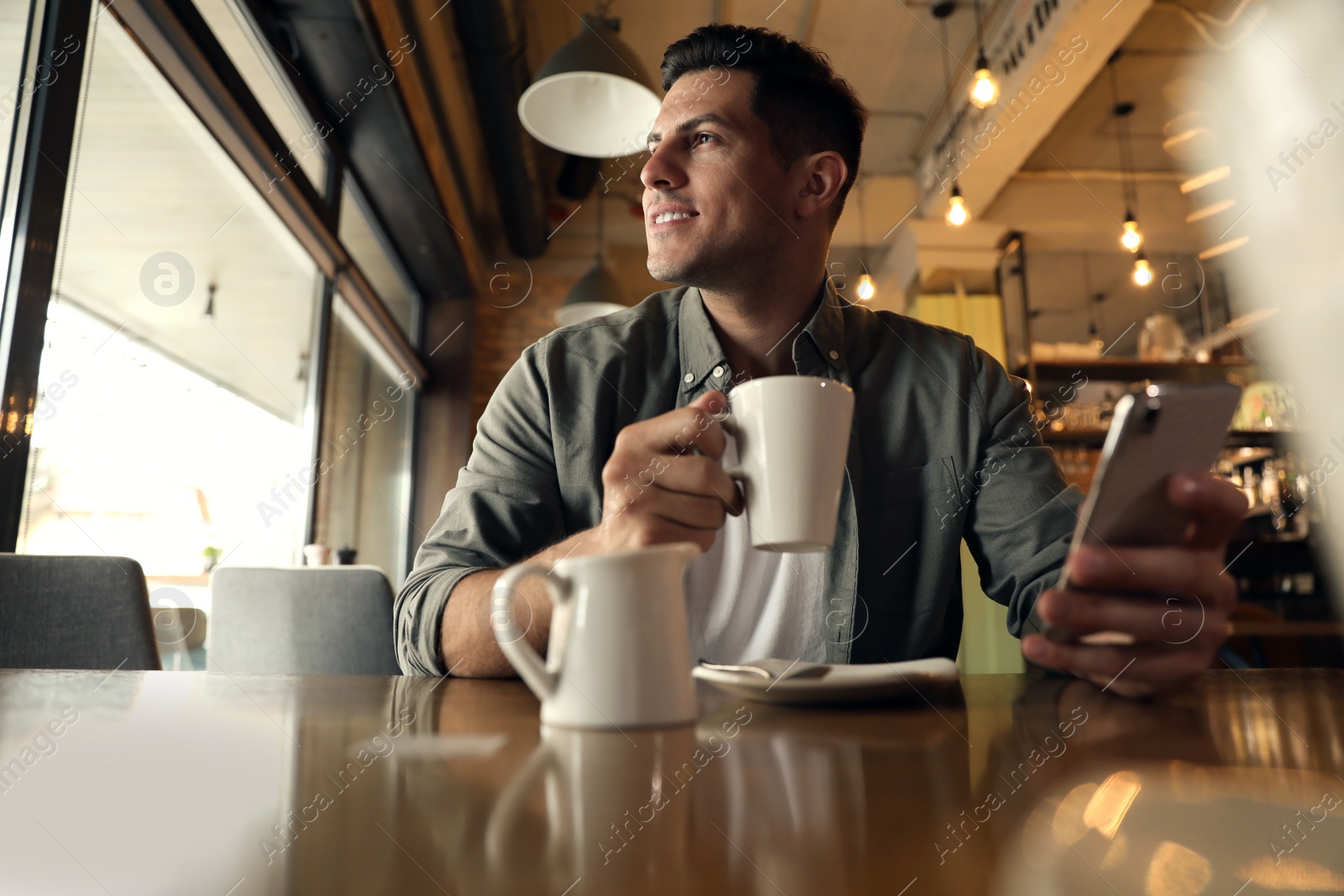 Photo of Handsome man with cup of coffee and smartphone at cafe in morning
