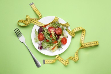 Photo of Plate with fresh vegetable salad, fork and measuring tape on light green background, flat lay. Healthy diet concept