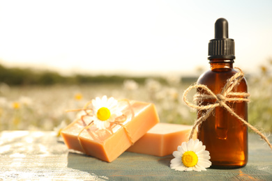 Photo of Bottle of chamomile essential oil and soap bars on blue wooden table in field