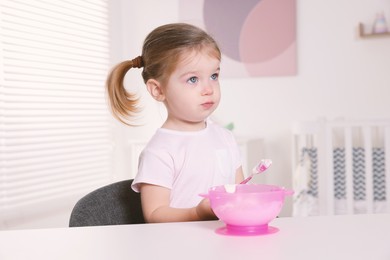 Photo of Cute little child eating tasty yogurt from plastic bowl with spoon at white table indoors