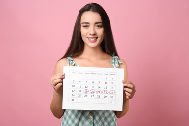 Young woman holding calendar with marked menstrual cycle days on pink background