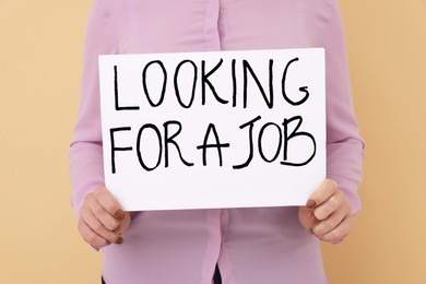 Photo of Young unemployed woman holding sign with phrase Looking For A Job on beige background, closeup