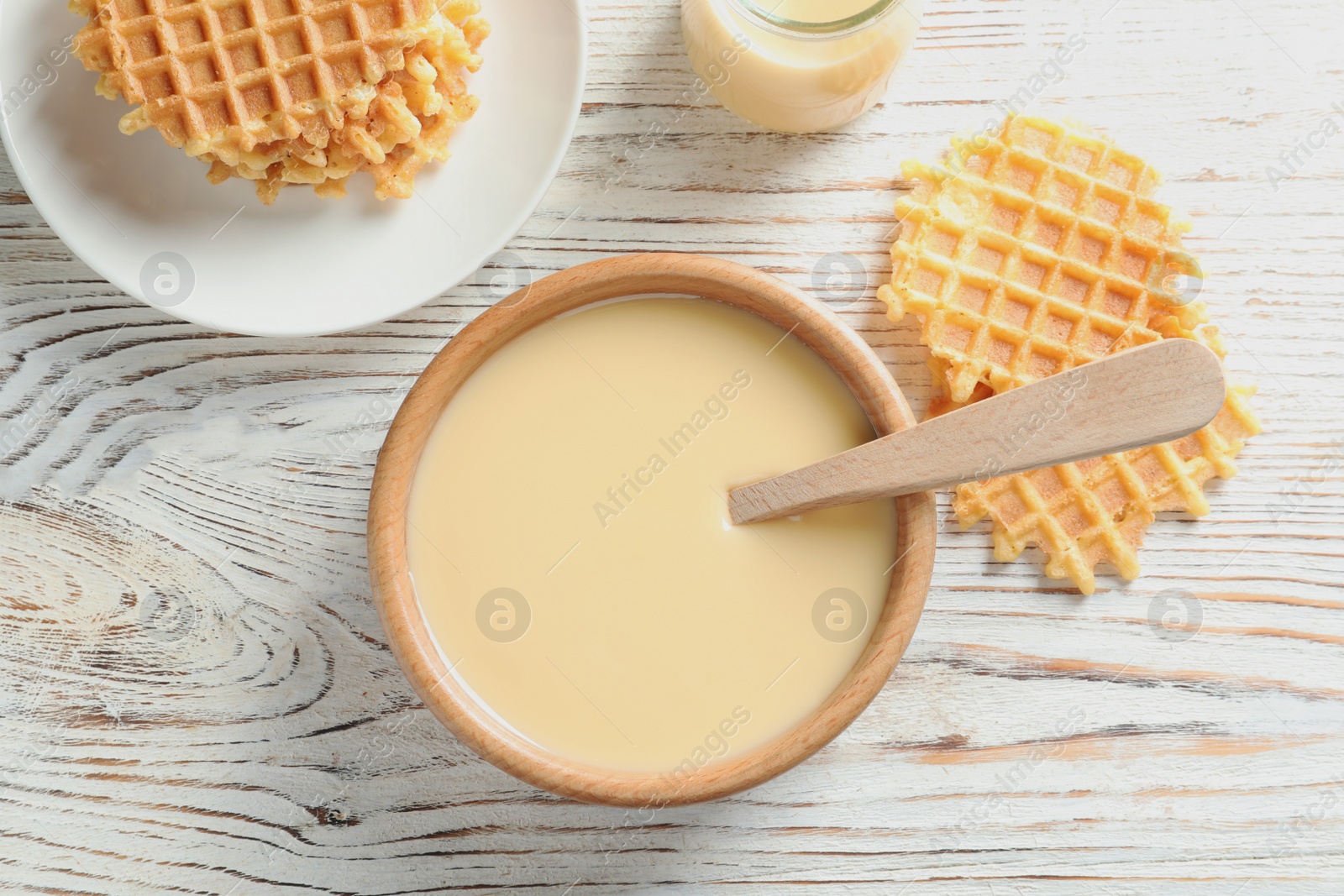 Photo of Bowl of condensed milk and waffles served on wooden table, top view. Dairy products