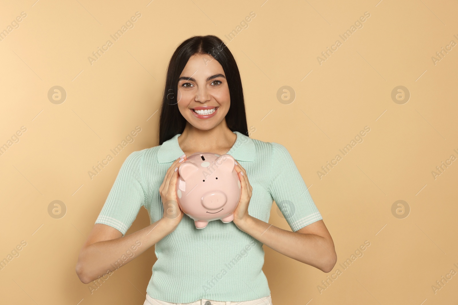 Photo of Happy young woman with ceramic piggy bank on beige background