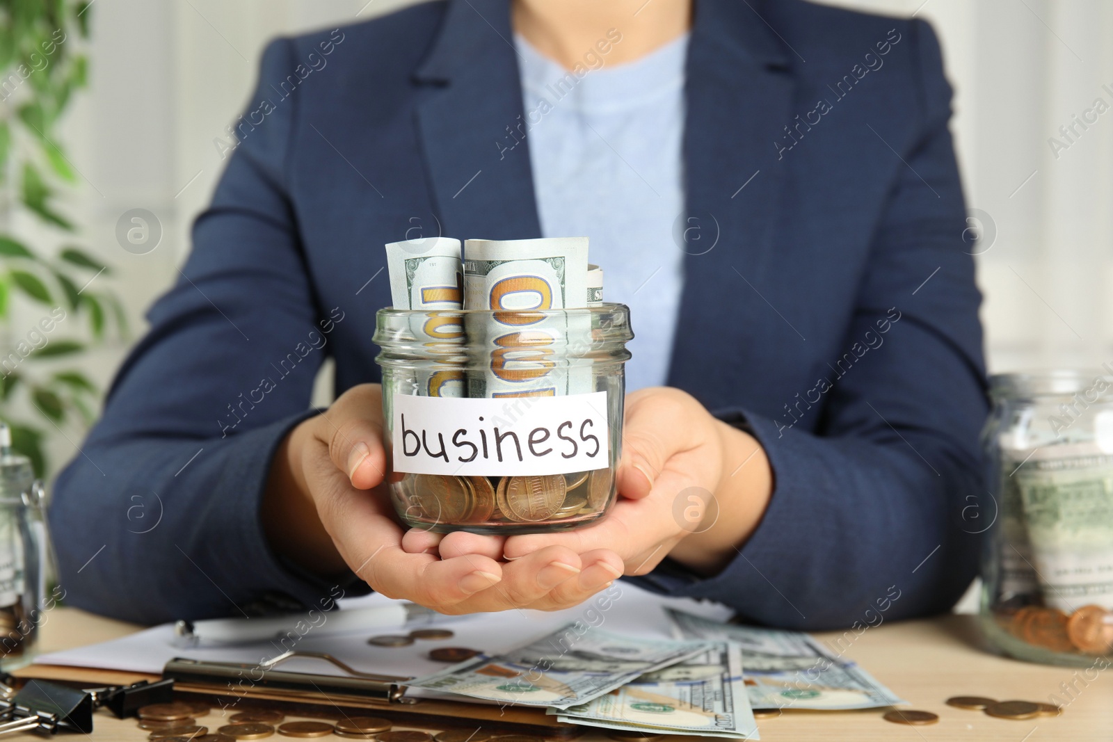 Photo of Woman holding glass jar with money and tag BUSINESS at wooden table, closeup