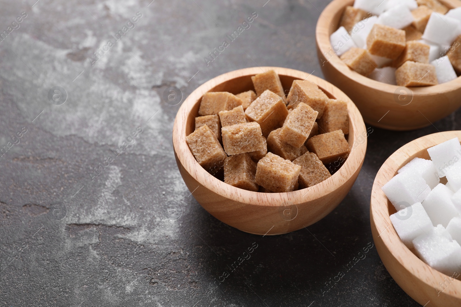 Photo of Different sugar cubes in bowls on gray textured table, closeup. Space for text