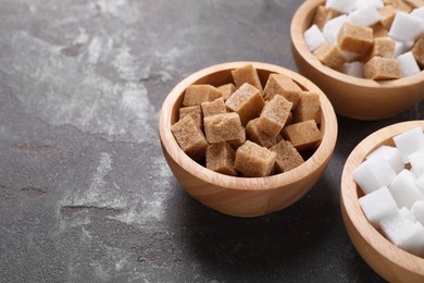 Different sugar cubes in bowls on gray textured table, closeup. Space for text