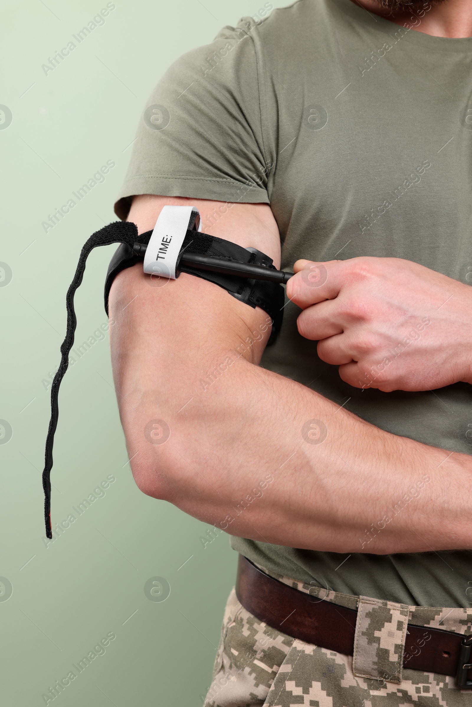 Photo of Soldier in military uniform applying medical tourniquet on arm against light olive background, closeup
