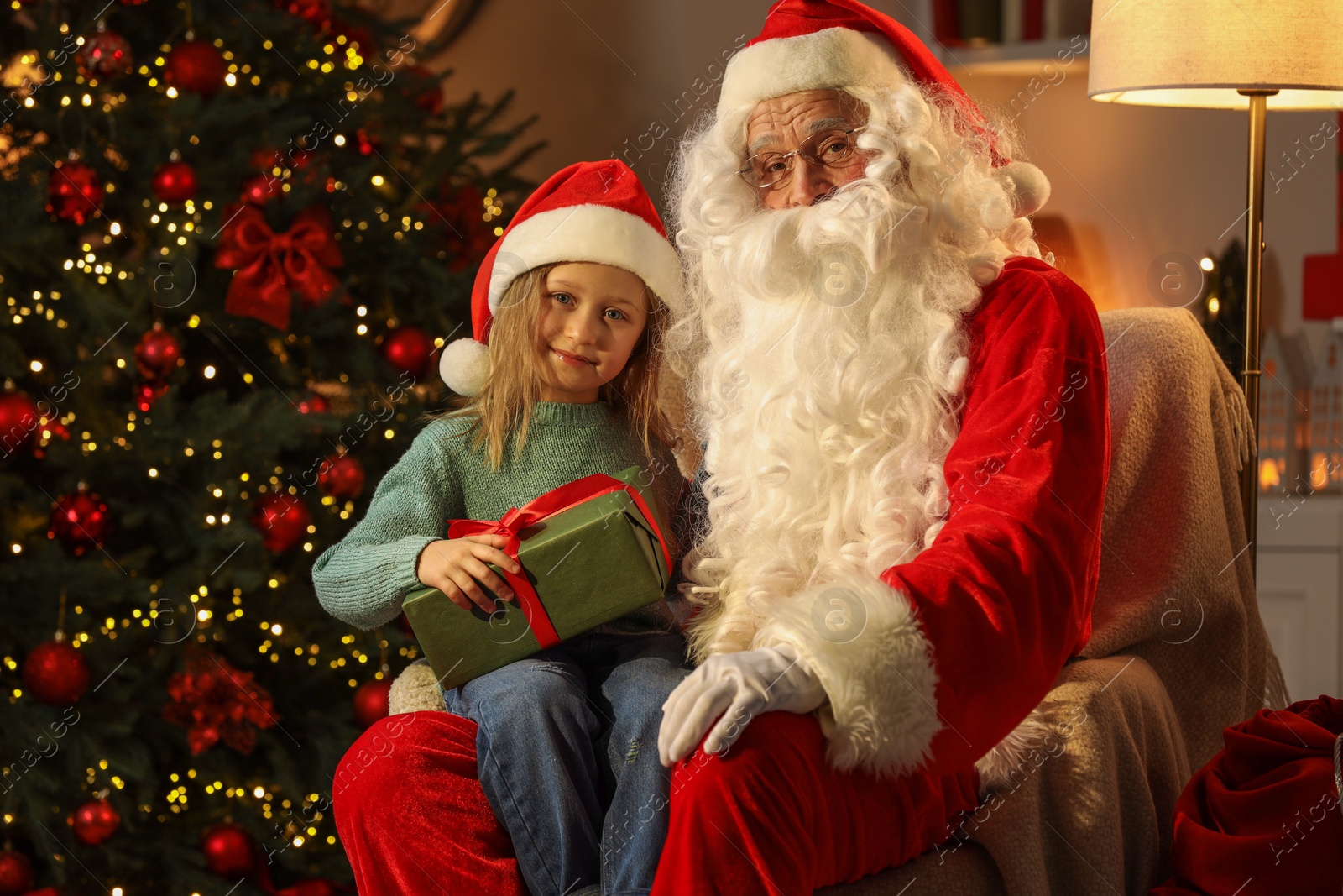 Photo of Merry Christmas. Little girl sitting on Santa's knee and holding gift at home