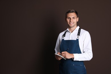 Portrait of happy young waiter with notebook on brown background, space for text
