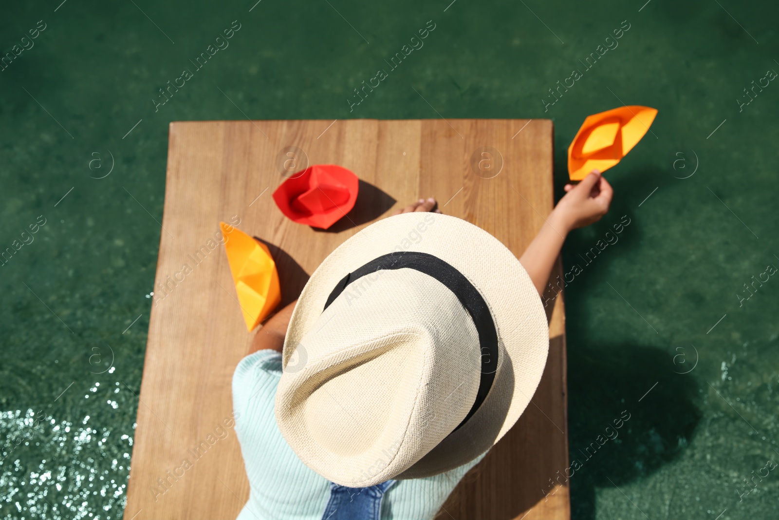 Photo of Little child playing with paper boat on wooden pier near river, above view