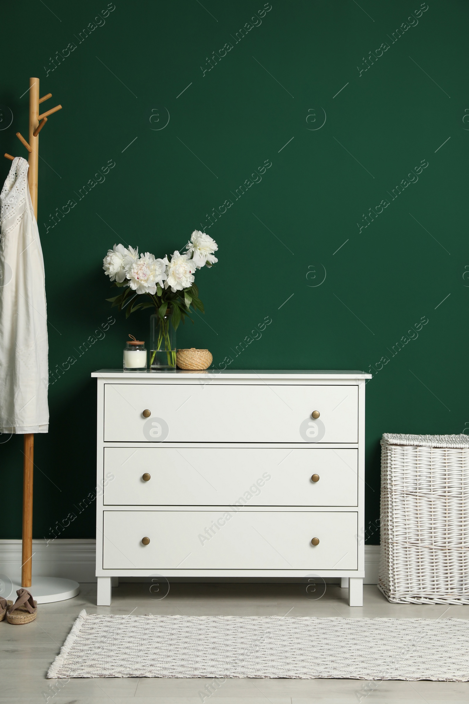 Photo of Modern white chest of drawers, coat stand and wicker basket near green wall indoors