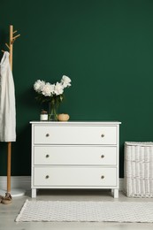Photo of Modern white chest of drawers, coat stand and wicker basket near green wall indoors