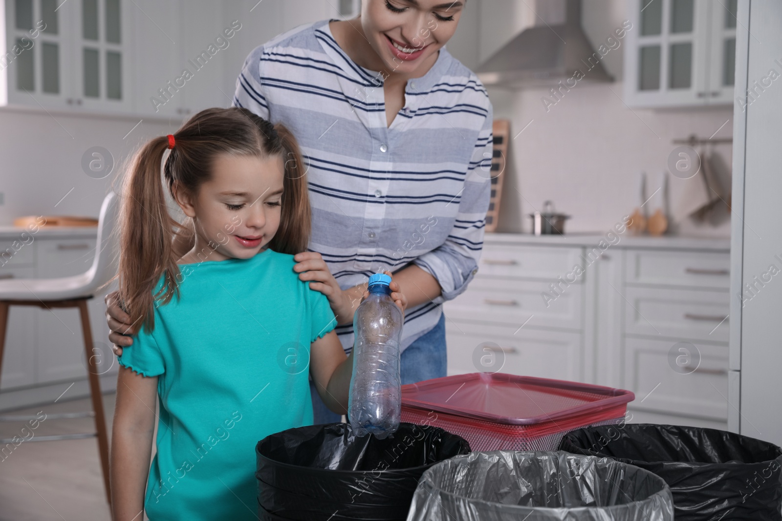 Photo of Young woman and her daughter throwing plastic bottle into trash bin in kitchen. Separate waste collection