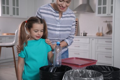 Photo of Young woman and her daughter throwing plastic bottle into trash bin in kitchen. Separate waste collection