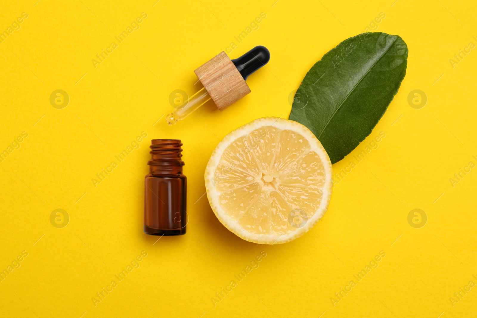 Photo of Bottle of citrus essential oil, pipette and fresh lemon on yellow background, flat lay