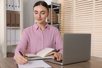 Photo of Woman taking notes while using laptop at wooden table in office