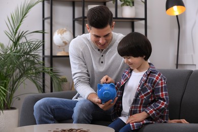 Photo of Little boy with his father putting coin into piggy bank at home