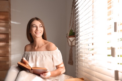 Beautiful young woman reading book near window at home