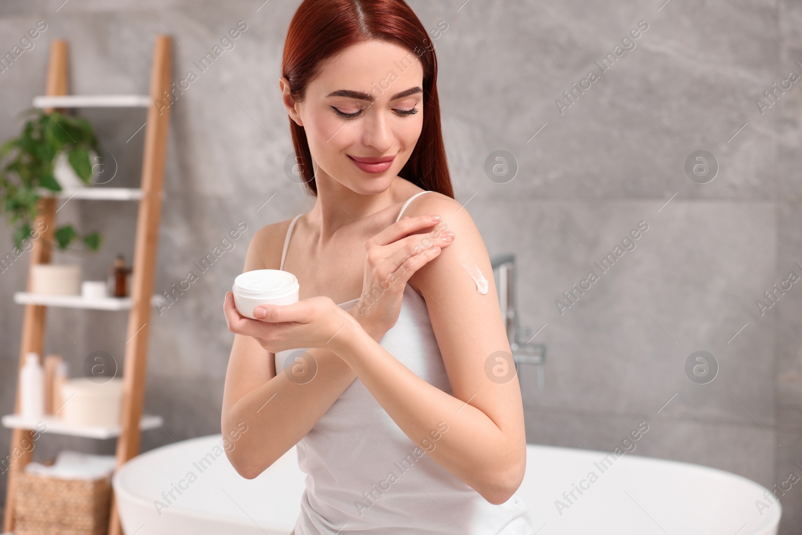 Photo of Beautiful young woman applying body cream onto shoulder in bathroom