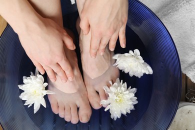 Photo of Woman soaking her feet in bowl with water and flowers on floor, top view. Spa treatment