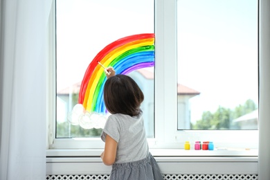 Photo of Little girl drawing rainbow on window indoors, back view. Stay at home concept