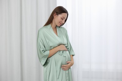 Beautiful pregnant woman in green dressing gown near window indoors