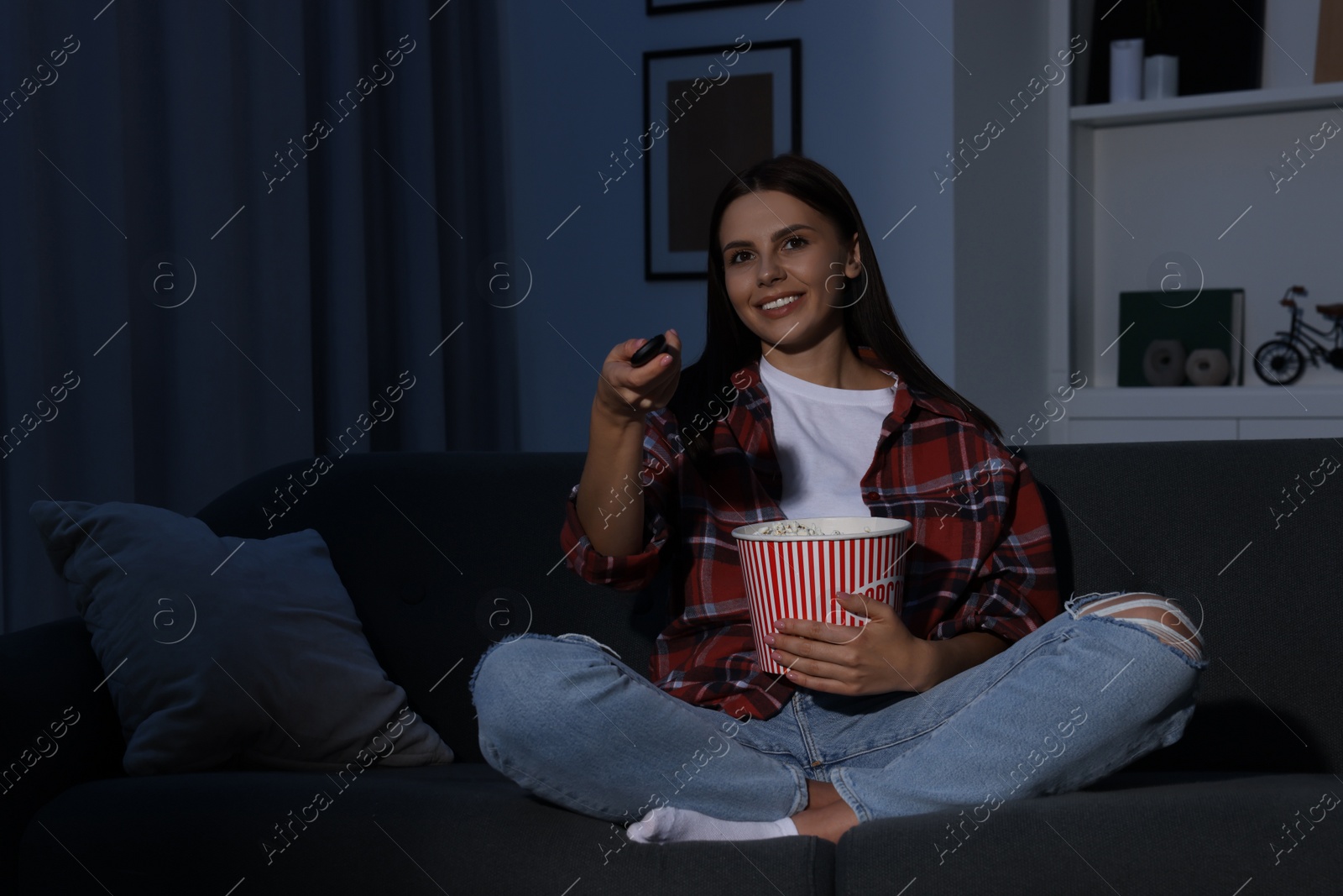 Photo of Happy woman holding popcorn bucket and changing TV channels with remote control at home in evening