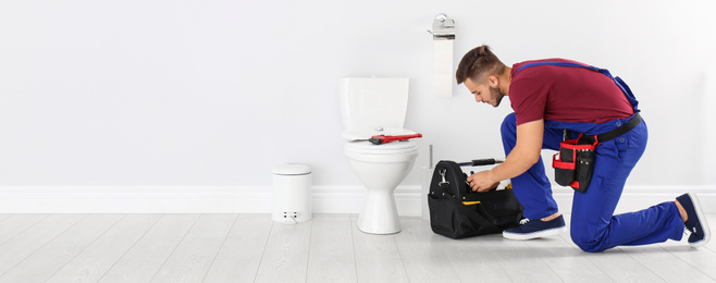 Young man with tool kit bag near toilet bowl in bathroom, space for text. Banner design